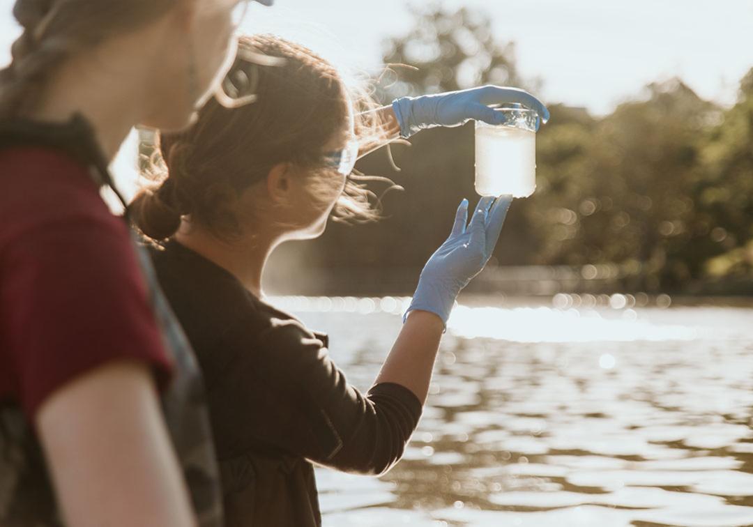 Two female students next to a river, one holding up a glass of the river water. 