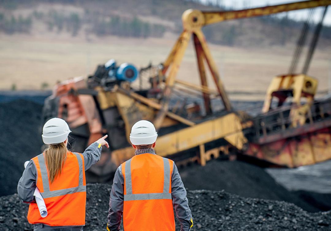 Two people walking on a mine site wearing high-vis and hard hats. Heavy machinery operates in the background.