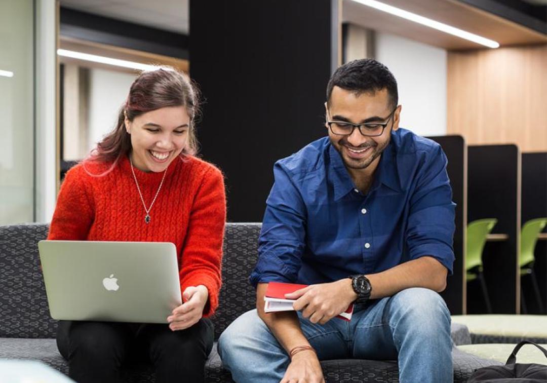 Two students sitting next to each other, one with a laptop. Both are smiling.