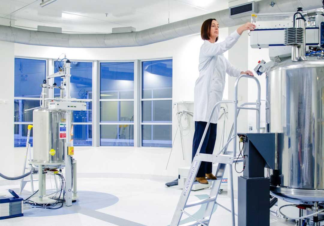 A UQ expert in a lab coat stands on a ladder getting readings from a large piece of equipment