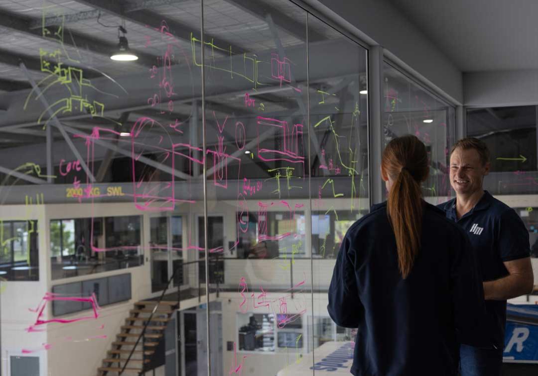 UQ expert works with a partner to discuss equations written on glass overlooking a workshop