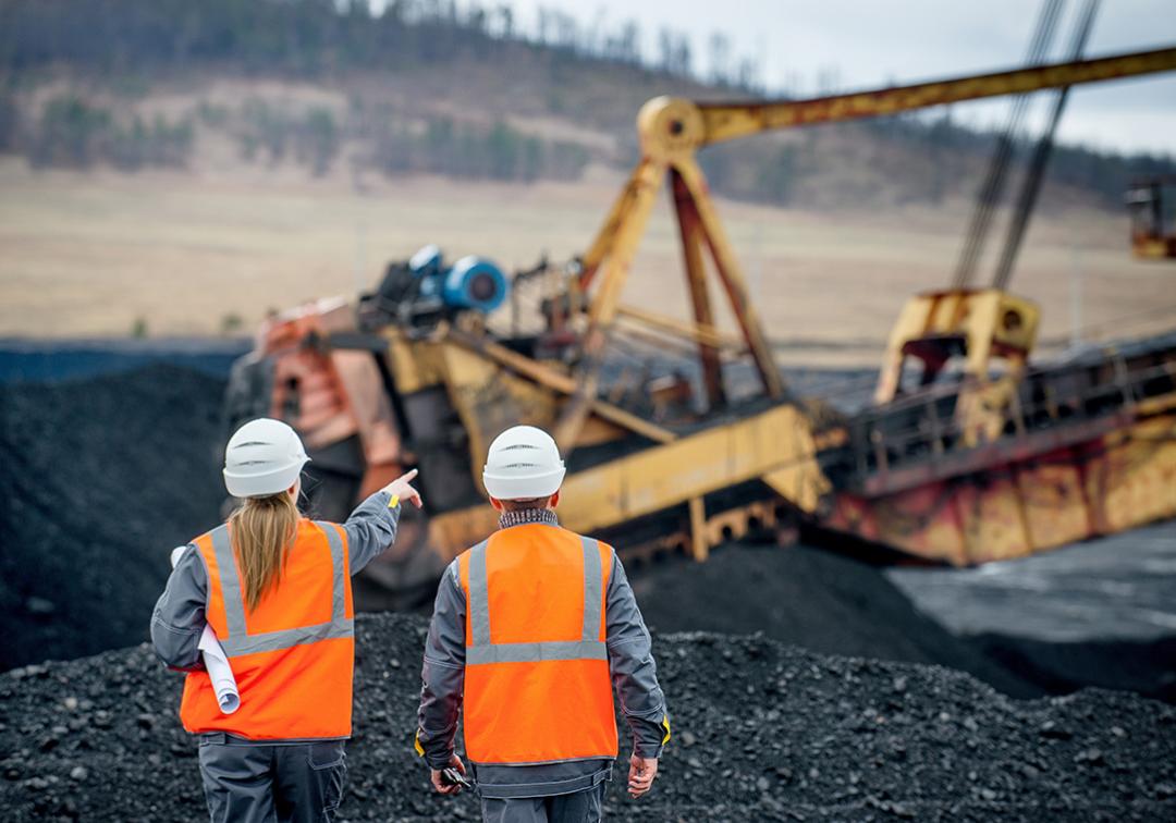 Two people walking on a mine site wearing high-vis and hard hats. Heavy machinery operates in the background.