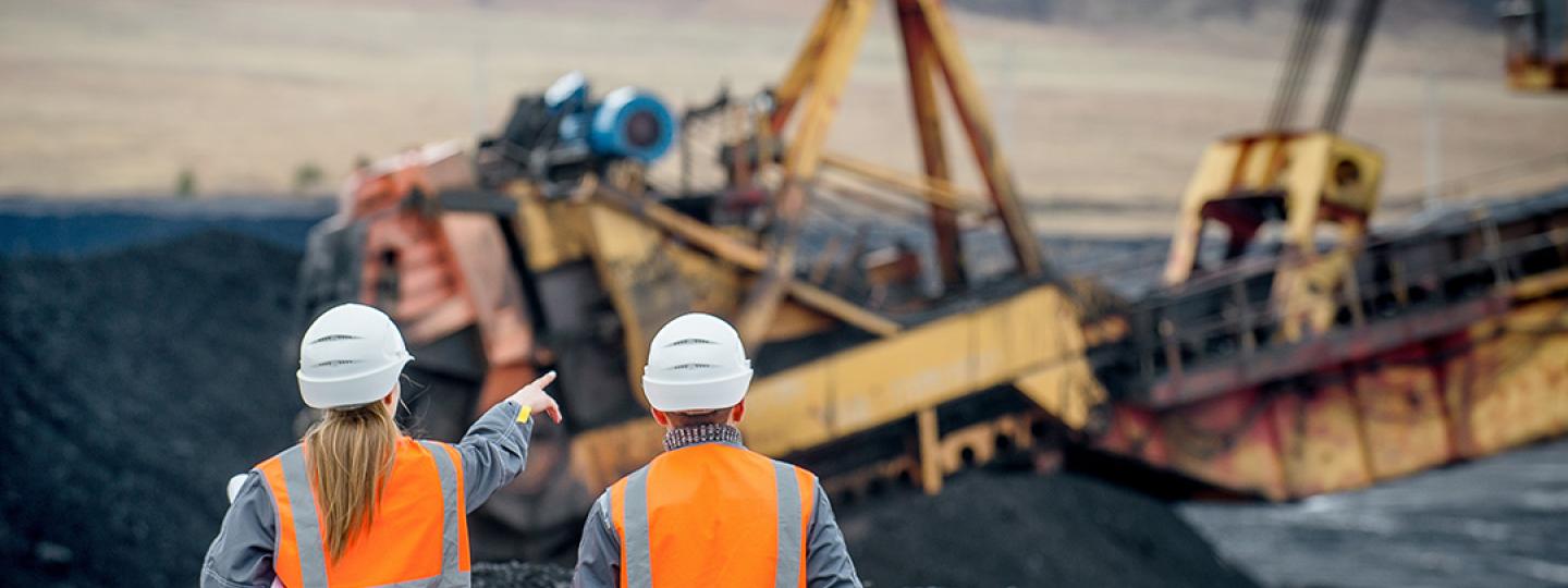 Two people walking on a mine site wearing high-vis and hard hats. Heavy machinery operates in the background.