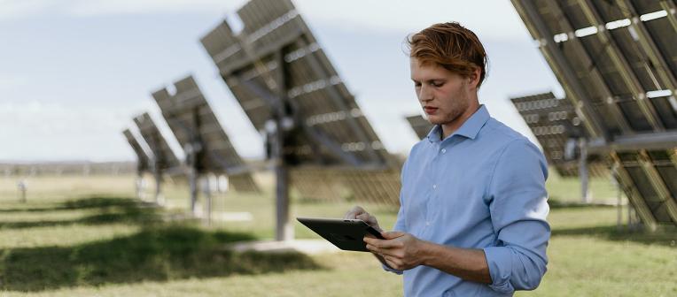 A man checks information on a tablet. Solar panels extend across a grassy field in the background.