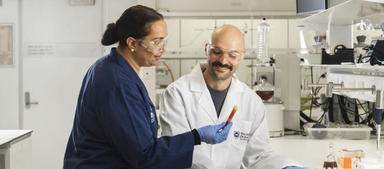 Two people wearing lab coats and safety glasses using equipment in a lab. 