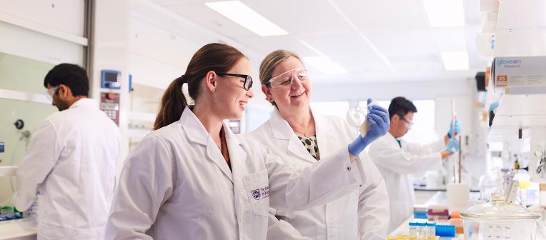 Two people wearing lab coats and safety glasses examining a test tube.