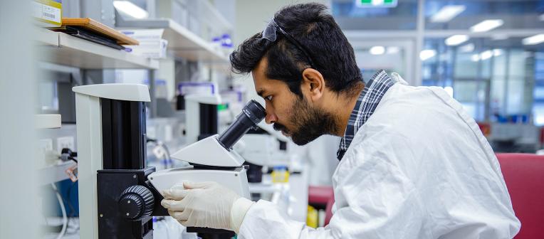 A man wearing a white lab coat and gloves looks into a microscope.