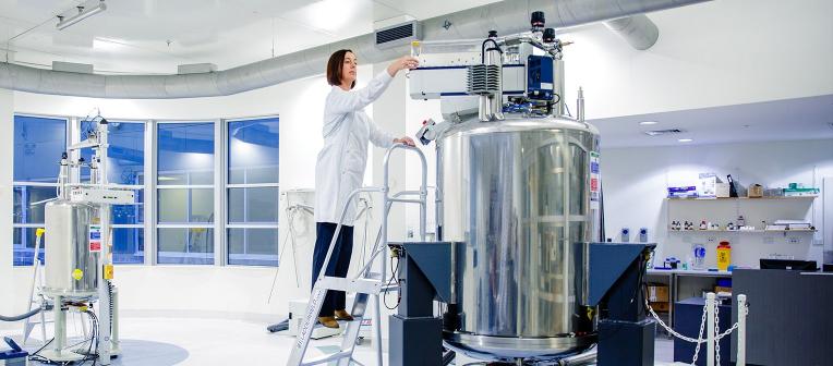 A woman wearing a lab coat standing on a step ladder, using equipment in a lab.