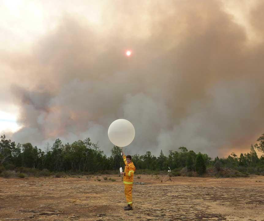A researcher with a large weather balloon stands in a dry field with bushfire smoke on the horizon