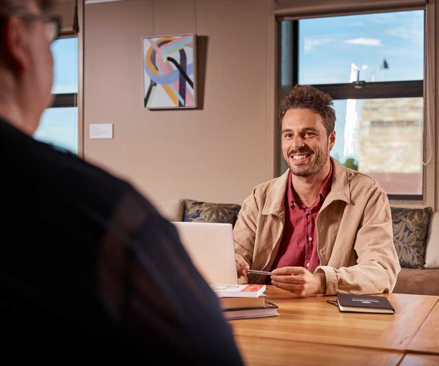 Two people discuss a project in an office with UQ's sandstone buildings visible in the background