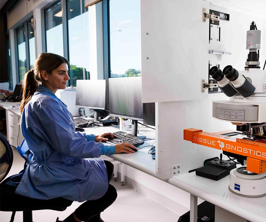 A woman wearing scrubs using a computer next to a piece of scientific equipment.