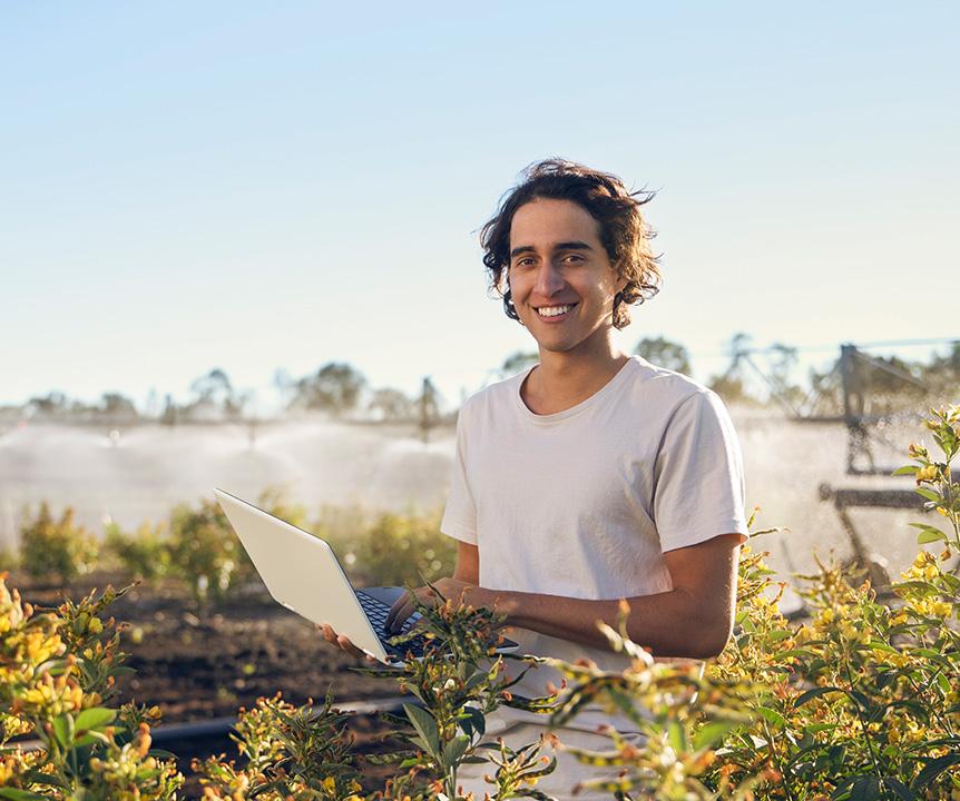 A person holding a laptop while standing in a field of crops. A large irrigation sprinkler operates in the background.