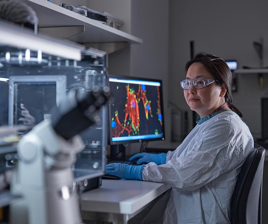 A woman wearing a lab coat and safety glasses working on a computer. A microscope sits in the foreground.