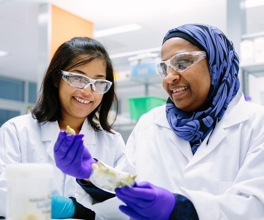 Two UQ researchers wearing white lab coats and safety glasses using equipment in a lab.