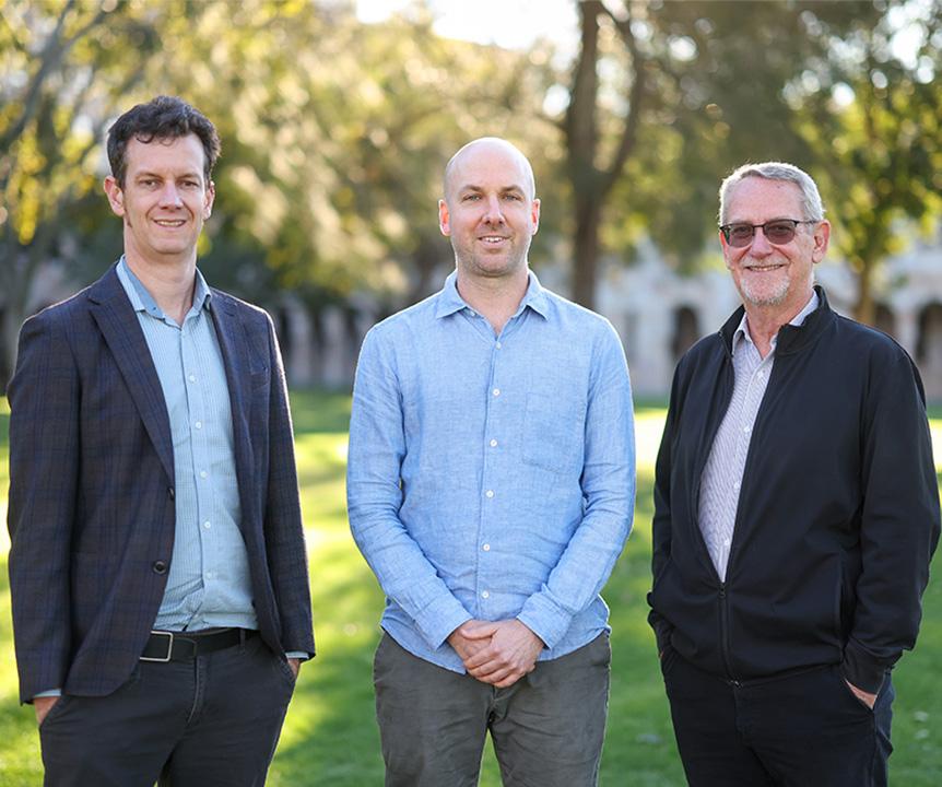 Three men stand in a grassy forecourt with sandstone buildings in the background.