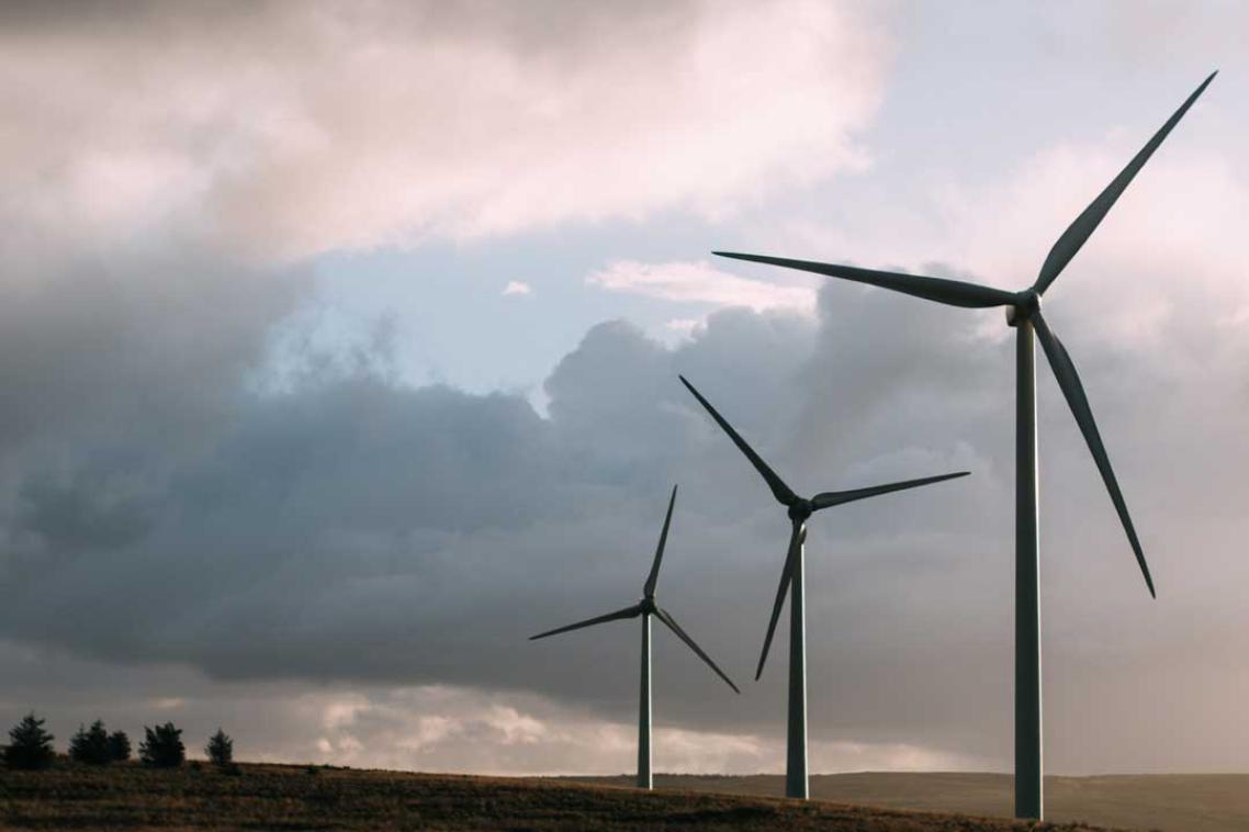 3 wind turbines spin on a grassy hillside with green trees in the background.