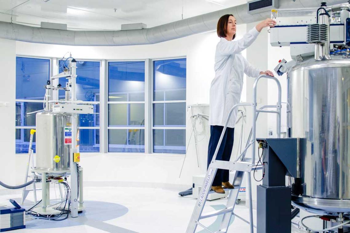 A UQ expert in a lab coat stands on a ladder getting readings from a large piece of equipment