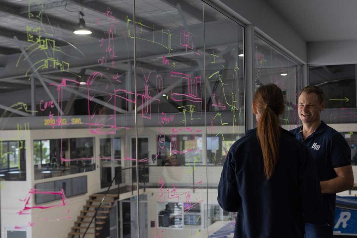 UQ expert works with a partner to discuss equations written on glass overlooking a workshop
