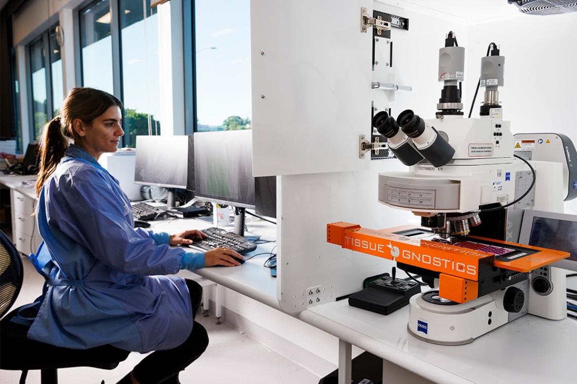 A woman wearing scrubs using a computer next to a piece of scientific equipment.