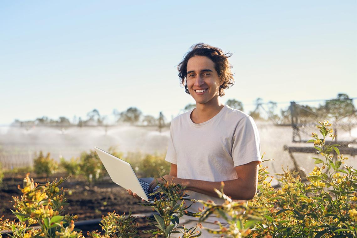 A man holding a laptop standing in a field of crops. A large irrigation sprinkler operates in the background.