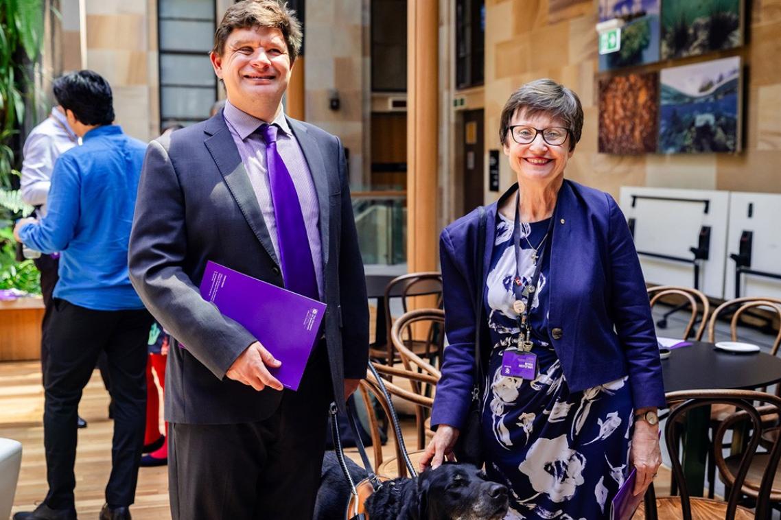 Two people standing together, one holding the harness handle of an assistance dog.